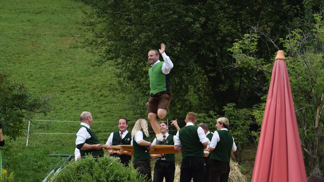 Eine Gruppe von Männern in traditionellen Trachten führt einen Sprung auf einer Holzplattform aus. Im Hintergrund ist eine grüne Landschaft mit Bäumen zu sehen. | © TVB Murau