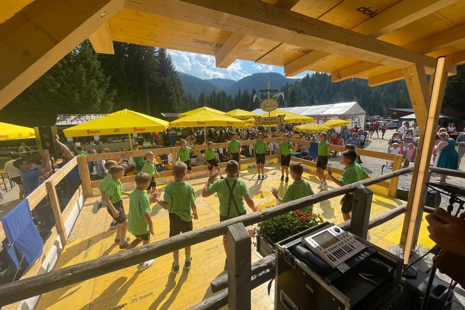 Eine Gruppe von Kindern in grünen T-Shirts tanzt auf einer Holzfläche. Im Hintergrund sind gelbe Sonnenschirme und Zuschauer zu sehen.