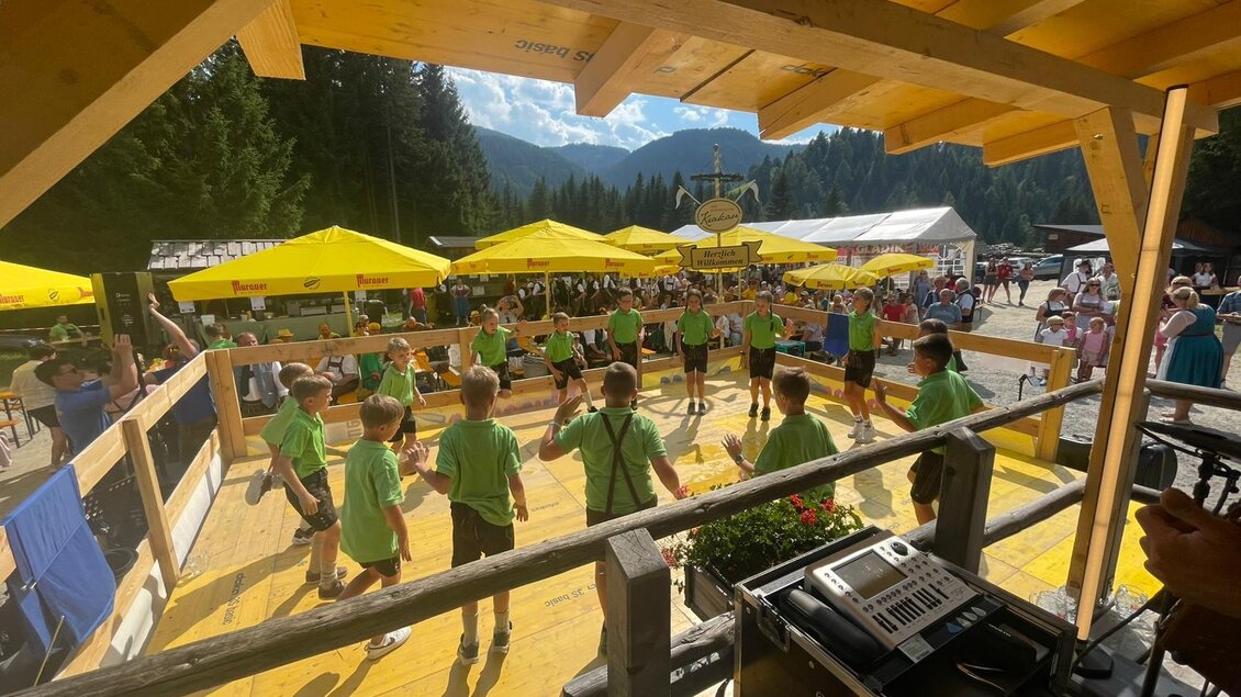 Eine Gruppe von Kindern in grünen T-Shirts tanzt auf einer Holzfläche. Im Hintergrund sind gelbe Sonnenschirme und Zuschauer zu sehen.