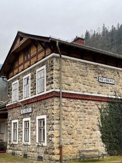 Ein historisches Bahnhofsgebäude aus Stein mit einem schönen Holzgiebel. Im Hintergrund sind bewaldete Berge zu sehen. | © Naturpark Mürzer Oberland