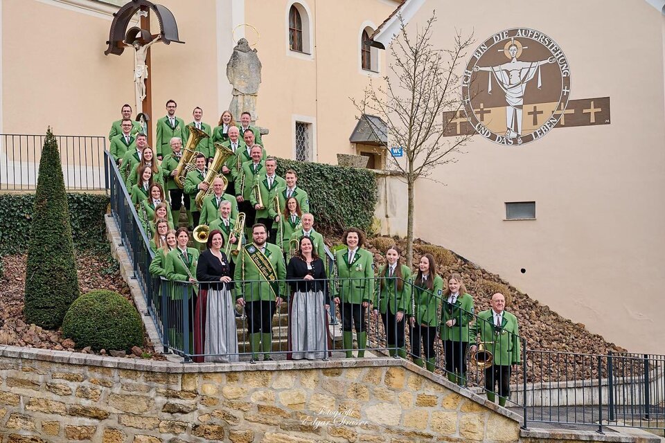 Eine Gruppe von Sängern in grünen Trachten steht auf einer Treppe vor einem Gebäude. Im Hintergrund ist ein religiöses Symbol zu sehen. | © Marktmusikkapelle St. Anna/Aigen