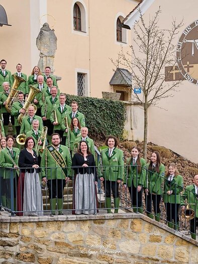 Eine Gruppe von Sängern in grünen Trachten steht auf einer Treppe vor einem Gebäude. Im Hintergrund ist ein religiöses Symbol zu sehen. | © Marktmusikkapelle St. Anna/Aigen