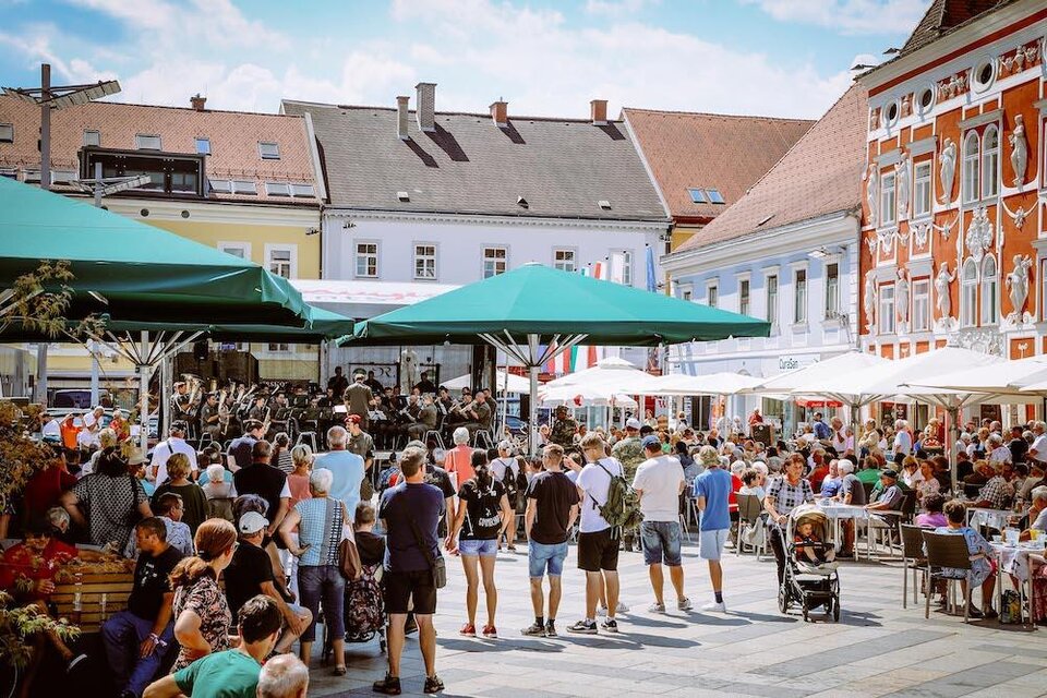 Ein belebter Platz mit vielen Menschen, die unter Sonnenschirmen sitzen. Im Hintergrund gibt es Stände und bunte Gebäude. | © Foto Freisinger