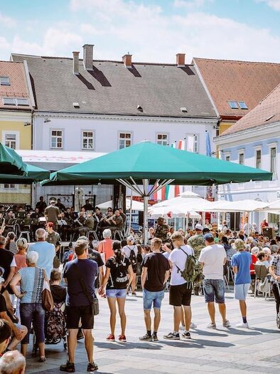 Ein belebter Platz mit vielen Menschen, die an einem Event teilnehmen. Bunte Gebäude und Sonnenschirme schaffen eine gemütliche Atmosphäre. | © Bild: Foto Freisinger