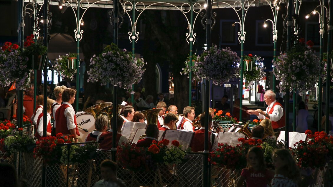 Eine Blasmusikgruppe spielt in einem festlich dekorierten Pavillon. Der Platz ist mit Blumen geschmückt und ist abends beleuchtet. | © Harald Steiner