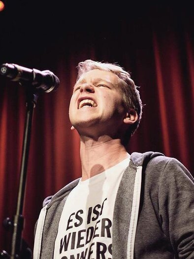 A man passionately sings into the microphone. In the background, there are red curtains that create a stage atmosphere. | © Marvin Ruppert