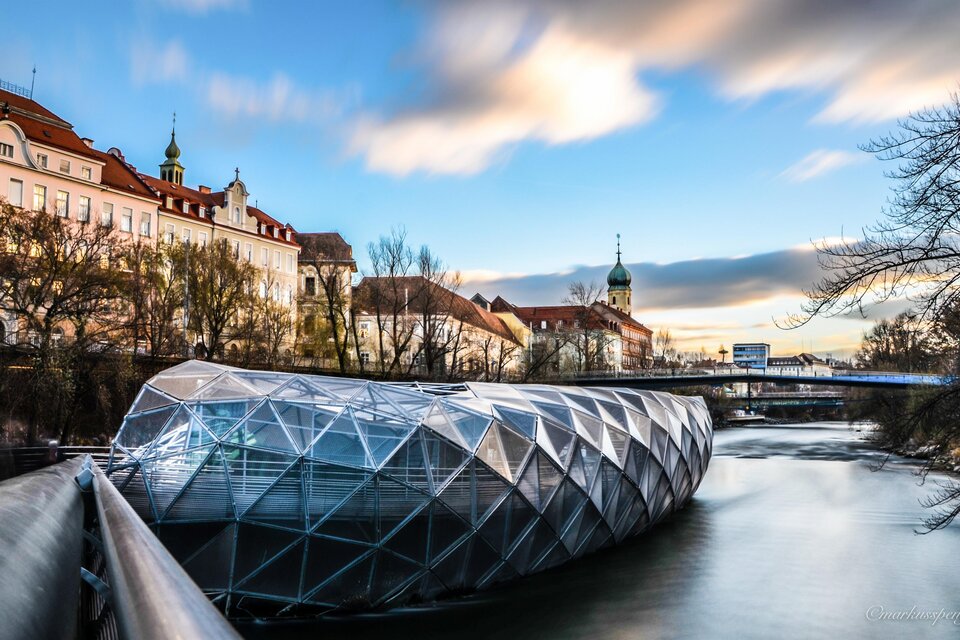 Eine moderne, gläserne Struktur über einem Fluss, umgeben von historischen Gebäuden. Der Himmel ist klar und die Umgebung ist ruhig und einladend. | © (c) Graz Tourismus - Markus Spenger