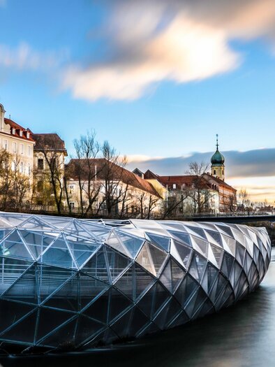 A modern, glass structure over a river, surrounded by historic buildings. The sky is clear and the surroundings are calm and inviting. | © (c) Graz Tourismus - Markus Spenger