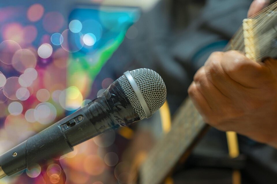 Ein Mikrofon steht im Vordergrund, während eine Hand eine Gitarre spielt. Der Hintergrund zeigt farbige Lichter, die eine festliche Atmosphäre schaffen. | © pixabay.com