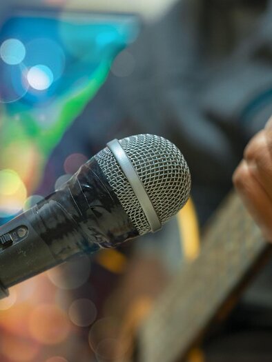 Ein Mikrofon steht im Vordergrund mit unscharfen bunten Lichtern im Hintergrund. Eine Hand spielt eine Gitarre neben dem Mikrofon. | © pixabay.com