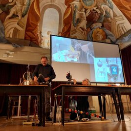 Eine Präsentation in einem historischen Raum mit aufwändiger Deckenmalerei. Zwei Personen stehen an Tischen mit wissenschaftlichen Geräten und einem Bildschirm im Hintergrund. | © Echophysics