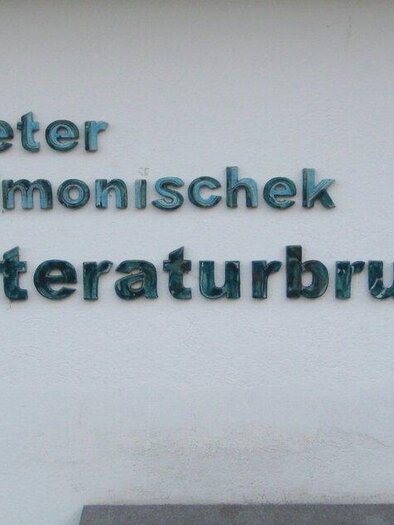 Eine Wand mit dem Schriftzug "peter simonischek literaturbrunnen". Darunter sind kunstvolle Gesichter mit Brillen und Gesichtszügen zu sehen. | © Gemeinde Markt Hartmannsdorf | Sarah Raiser