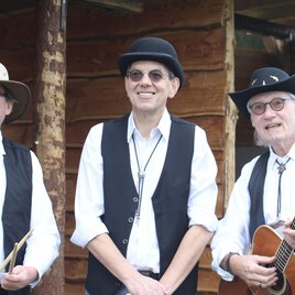 Drei Musiker in traditioneller Kleidung stehen vor einer Holzstruktur. Einer von ihnen hält eine Gitarre, während die anderen Musikinstrumente bei sich haben. | © Oststeiermark Tourismus