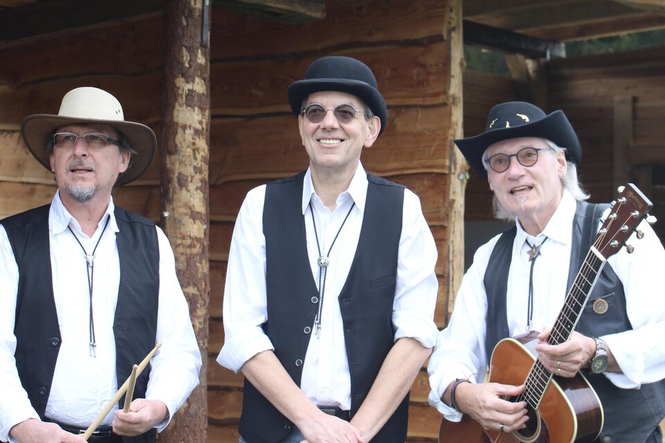 Drei Musiker in traditioneller Kleidung stehen vor einer Holzstruktur. Einer von ihnen hält eine Gitarre, während die anderen Musikinstrumente bei sich haben. | © Oststeiermark Tourismus
