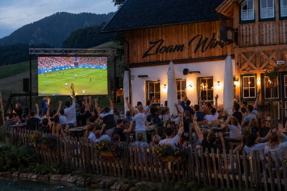 Eine lebhafte Außenveranstaltung mit einer großen Menschenmenge, die ein Fußballspiel auf einem Bildschirm verfolgt. Die Zuschauer jubeln begeistert und befinden sich in einer gemütlichen Umgebung.