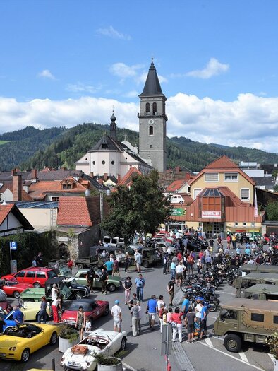 Ein farbenfrohes Stadtfest mit einer großen Ansammlung von Autos und Motorrädern. Im Hintergrund sieht man eine Kirche und grüne Hügel unter einem klaren blauen Himmel. | © Foto Mitteregger