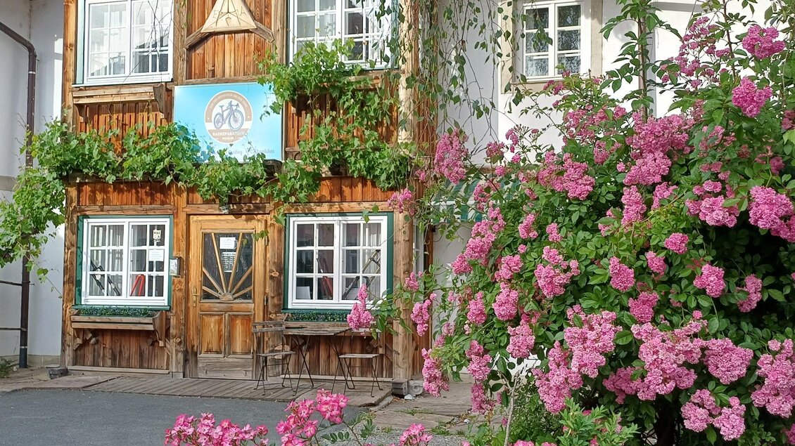 Ein charmantes Holzgebäude mit Fenstern und Pflanzen. Vor dem Haus blühen schöne rosa Blumen. | © TV Südsteiermark - Irene Löschnig