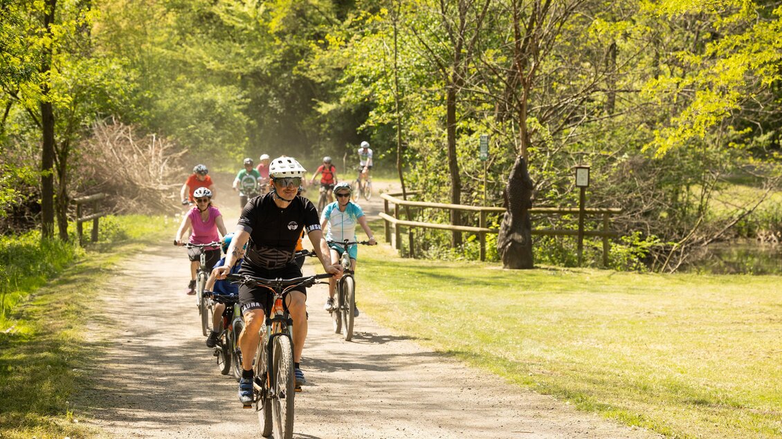 Eine Gruppe von Radfahrern fährt auf einem Schotterweg durch eine grüne Landschaft. Die Bäume sind in vollem Blätterkleid und die Sonne scheint. | © ZEHNERHAUS Bad Radkersburg