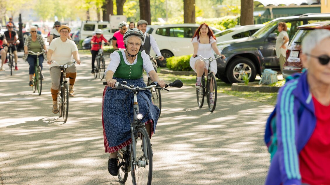Eine Gruppe von Radfahrern, darunter Frauen in traditionellen Kleidern, fährt an einer sonnigen Straße entlang. Bäume und Autos umgeben den Weg, was eine entspannte Atmosphäre schafft. | © ZEHNERHAUS Bad Radkersburg