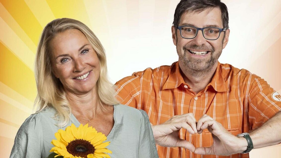 Ein lächelndes Paar steht vor einem sonnigen Hintergrund. Die Frau hält eine Sonnenblume und der Mann formt mit seinen Händen ein Herz. | © ORF/Regine Schöttl