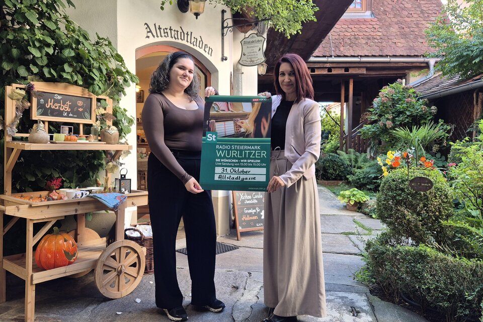 Zwei Frauen präsentieren vor dem liebevoll herbstlich dekorierten Altstadtladen in Feldbach ein Plakat für den Radio Steiermark Wurlitzer. Das stimmungsvolle Ambiente mit Kürbis, Blumen und viel Grün vermittelt gemütliches Herbstflair. | © Altstadtladen Feldbach