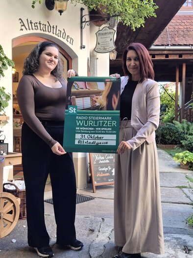 Two women are presenting a poster for the Radio Steiermark Wurlitzer in front of the lovingly autumnally decorated old town shop in Feldbach. The atmospheric setting with pumpkins, flowers, and lots of greenery conveys a cozy autumn flair. | © Altstadtladen Feldbach
