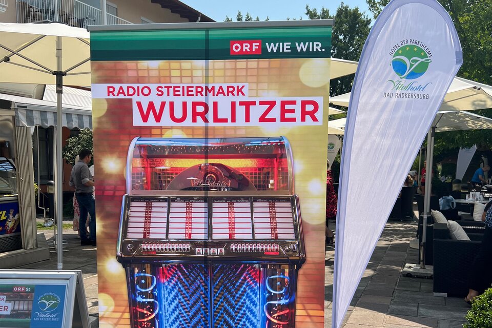 Ein buntes Werbebanner zeigt eine Wurlitzer-Jukebox. Im Hintergrund sind Sonnenschirme und eine Veranstaltung im Freien zu sehen. | © Vitalhotel der Parktherme