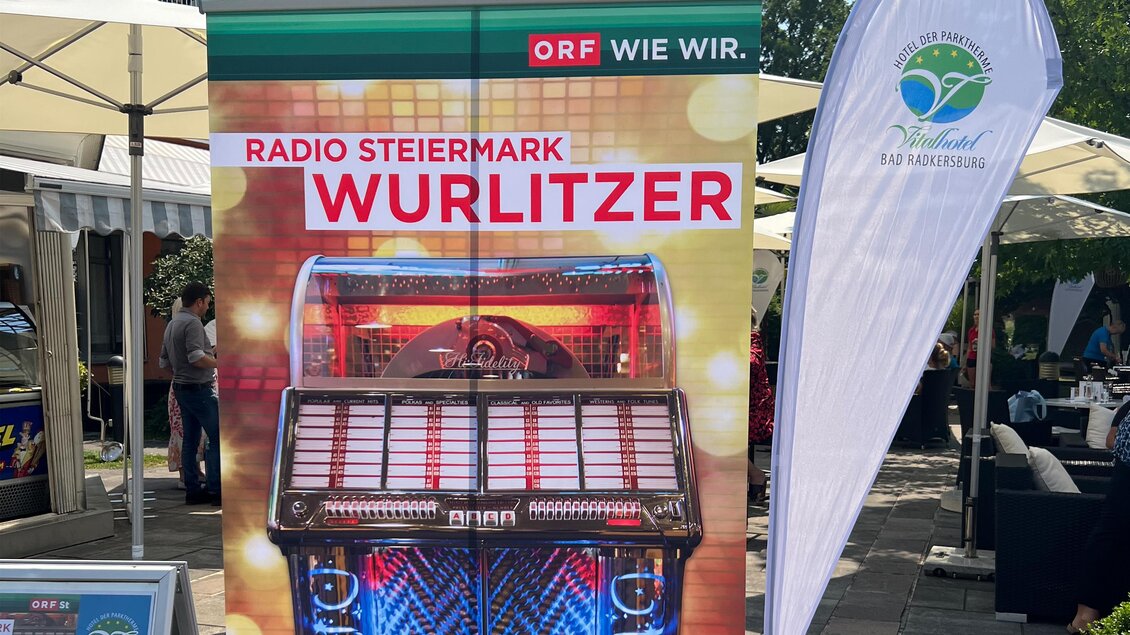 Ein buntes Werbebanner zeigt eine Wurlitzer-Jukebox. Im Hintergrund sind Sonnenschirme und eine Veranstaltung im Freien zu sehen. | © Vitalhotel der Parktherme