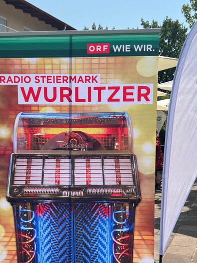 Ein buntes Werbebanner zeigt eine Wurlitzer-Jukebox. Im Hintergrund sind Sonnenschirme und eine Veranstaltung im Freien zu sehen. | © Vitalhotel der Parktherme