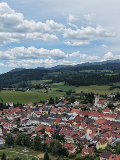 Eine malerische Stadt mit bunten Häusern, umgeben von grünen Feldern und Hügeln. Der Himmel ist teilweise bewölkt und die Landschaft wirkt einladend. | © TV Murau