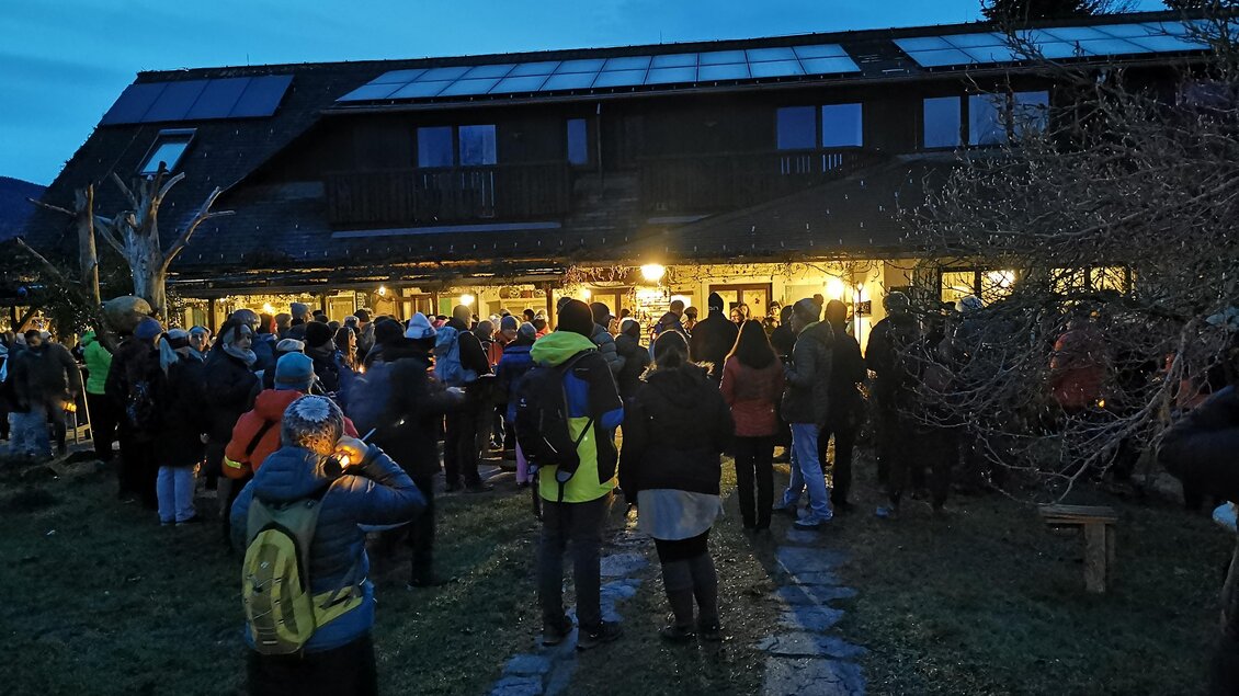 Eine Gruppe von Menschen versammelt sich vor einem Gebäude bei Dämmerung. Im Hintergrund leuchten die Lichter des Hauses. | © CDM