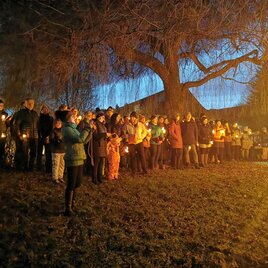 A group of people stands beside a large tree at dusk. Many are holding candles in their hands, creating a warm, festive atmosphere. | © cdm - Claudia Dunst