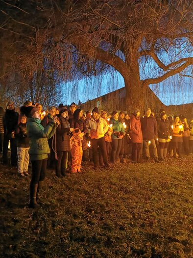 Eine Gruppe von Menschen steht bei Dämmerung neben einem großen Baum. Viele halten Kerzen in der Hand und schaffen eine warme, festliche Atmosphäre. | © cdm - Claudia Dunst