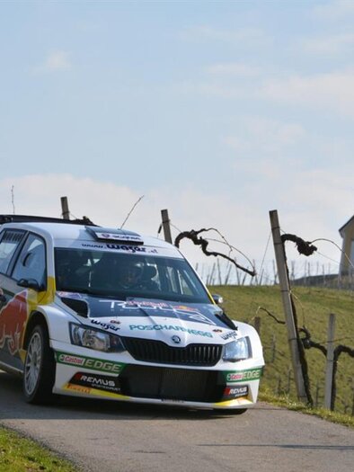 A racing car drives along a winding road between vineyards. In the background, a small church can be seen. | © Harald Illmer