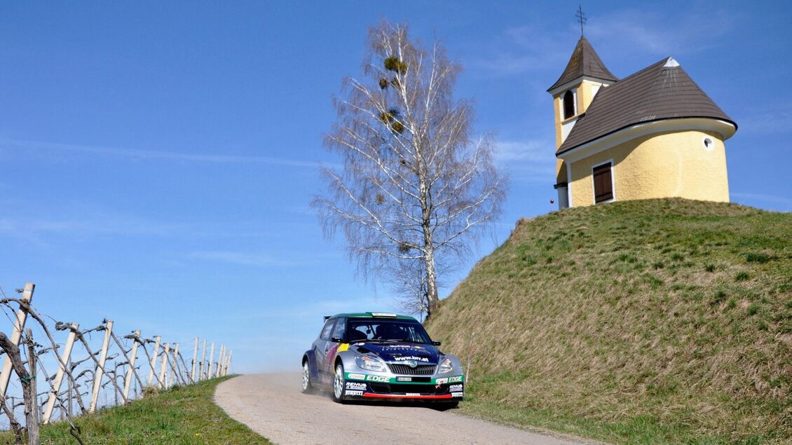 Ein Rallye-Auto fährt eine kurvenreiche Straße entlang eines Hügels. Im Hintergrund ist eine kleine Kirche und ein klarer blauer Himmel zu sehen. | © Harald Illmer
