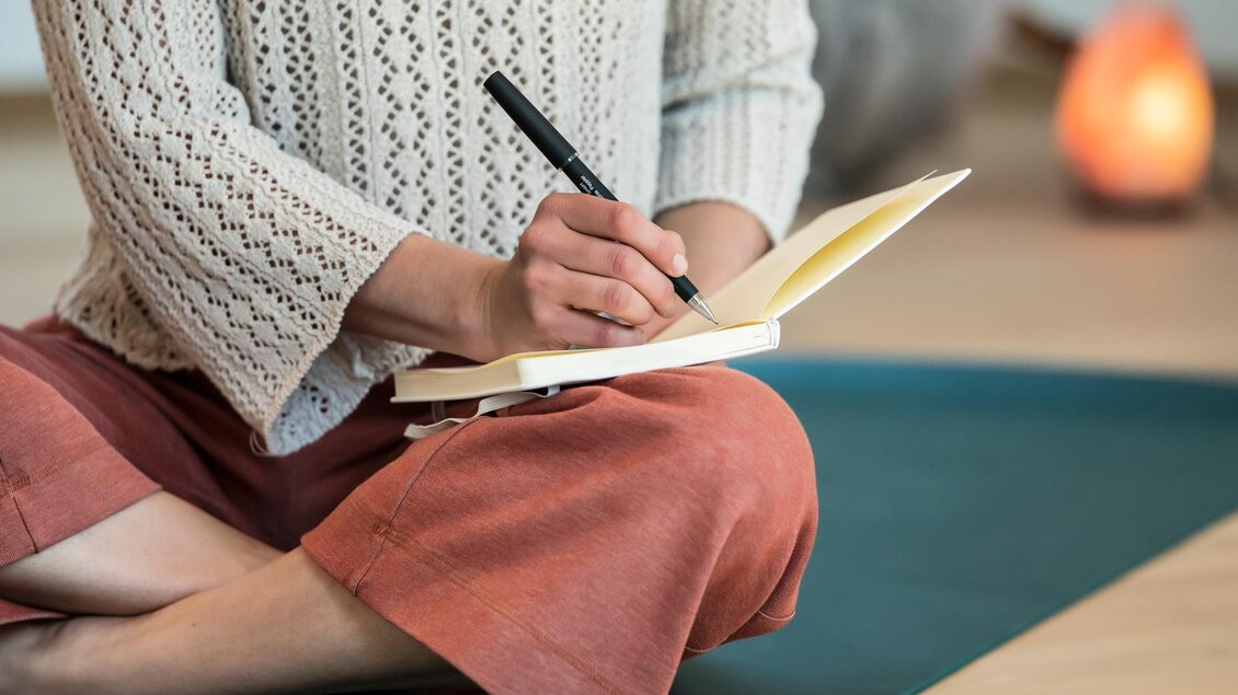 Eine Person sitzt auf einer Yogamatte und schreibt in ein Notizbuch. Im Hintergrund ist ein sanft leuchtendes Licht zu sehen.