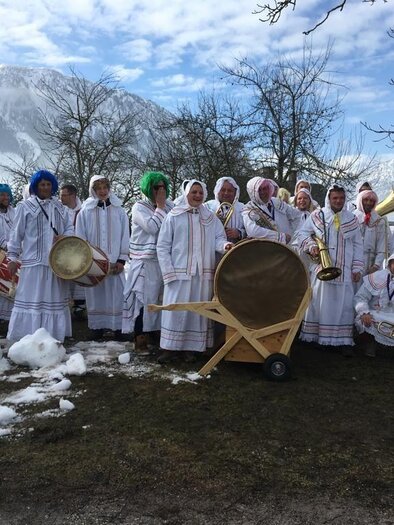 A group of people in traditional white clothing stands in the snow. They hold musical instruments and pose in front of a picturesque mountain landscape. | © Reinhard Pressl