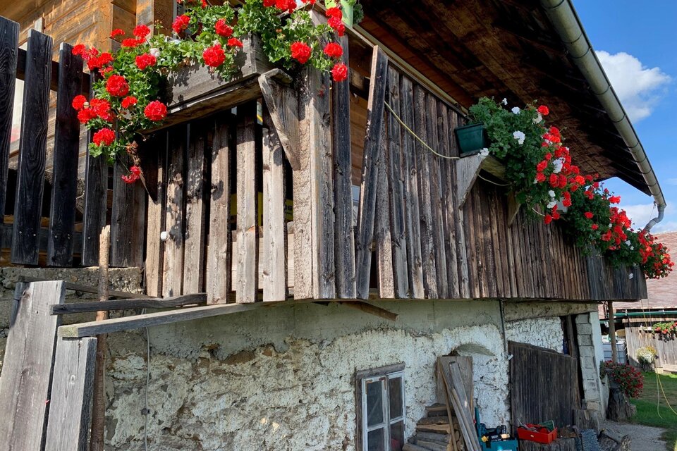 Ein traditionelles Haus mit einem Holzbalcon, der mit roten Blumen dekoriert ist. Die Fassade ist aus Stein und Holz, im Hintergrund ist ein blauer Himmel zu sehen. | © TV Südsteiermark