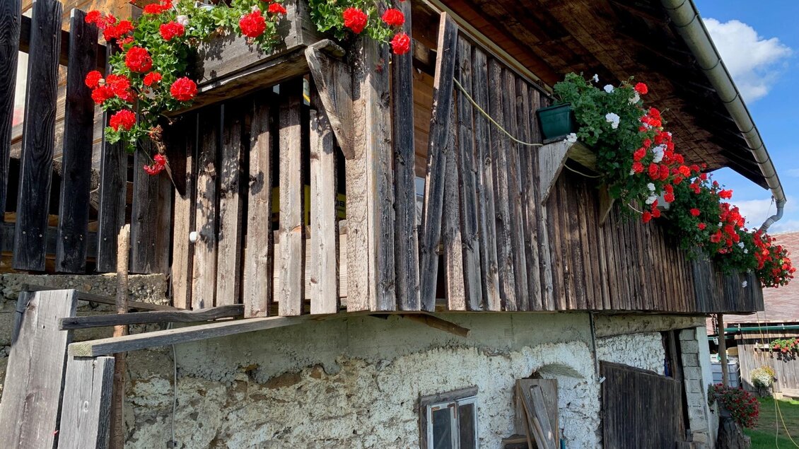 Ein traditionelles Haus mit einem Holzbalcon, der mit roten Blumen dekoriert ist. Die Fassade ist aus Stein und Holz, im Hintergrund ist ein blauer Himmel zu sehen. | © TV Südsteiermark