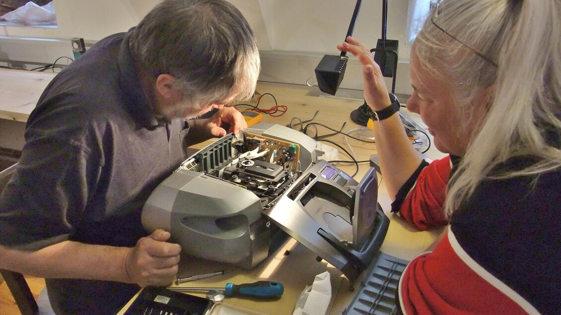Eine Person repariert eine Schreibmaschine, während eine andere zuschaut und diskutiert. Werkzeuge und Ersatzteile sind auf dem Tisch verteilt. | © Arzberger
