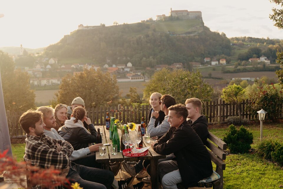 Eine Gruppe von Personen sitzt an einem Tisch im Freien und genießt die Aussicht auf eine Landschaft mit einem Hügel und einer Burg im Hintergrund. Es ist Abend und die Sonne scheint sanft. | © Dietmar Kump