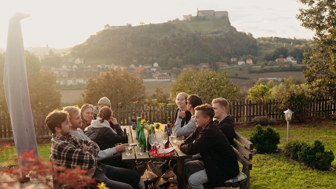 Eine Gruppe von Personen sitzt an einem Tisch im Freien und genießt die Aussicht auf eine Landschaft mit einem Hügel und einer Burg im Hintergrund. Es ist Abend und die Sonne scheint sanft. | © Dietmar Kump