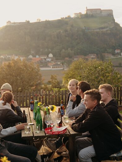 A group of people is sitting at a table outdoors, enjoying the view of a landscape with a hill and a castle in the background. It is evening, and the sun is shining gently. | © Dietmar Kump
