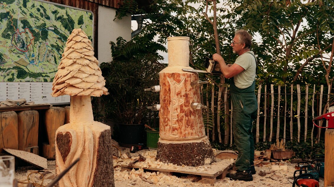 Ein Holzschnitzer arbeitet an einer Holzskulptur in einem Garten. Im Hintergrund sind weitere geschnitzte Figuren und eine Karte zu sehen. | © Dietmar Kump