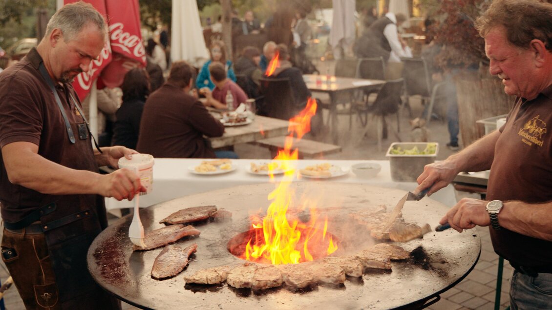 Zwei Männer grillen Fleisch über offenem Feuer auf einem großen Grill. Im Hintergrund sind Gäste, die das Essen genießen. | © Dietmar Kump