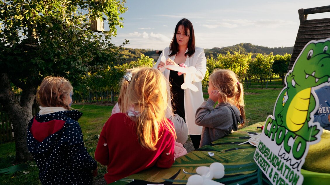 Eine Frau in einem weißen Kittel erklärt etwas Kindern im Freien. Im Hintergrund sind Weinreben und eine Naturlandschaft zu sehen. | © Dietmar Kump
