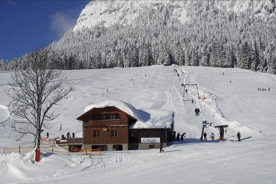 Eine verschneite Alpenlandschaft mit einem Holzhaus im Vordergrund. Im Hintergrund sind Skifahrer und ein Skilift zu sehen. | © Grafenwiese, Familie Sölkner