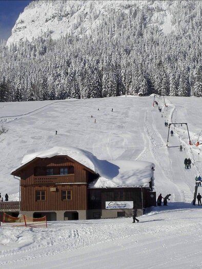 A snow-covered Alpine landscape with a wooden house in the foreground. In the background, skiers and a ski lift can be seen. | © Grafenwiese, Familie Sölkner