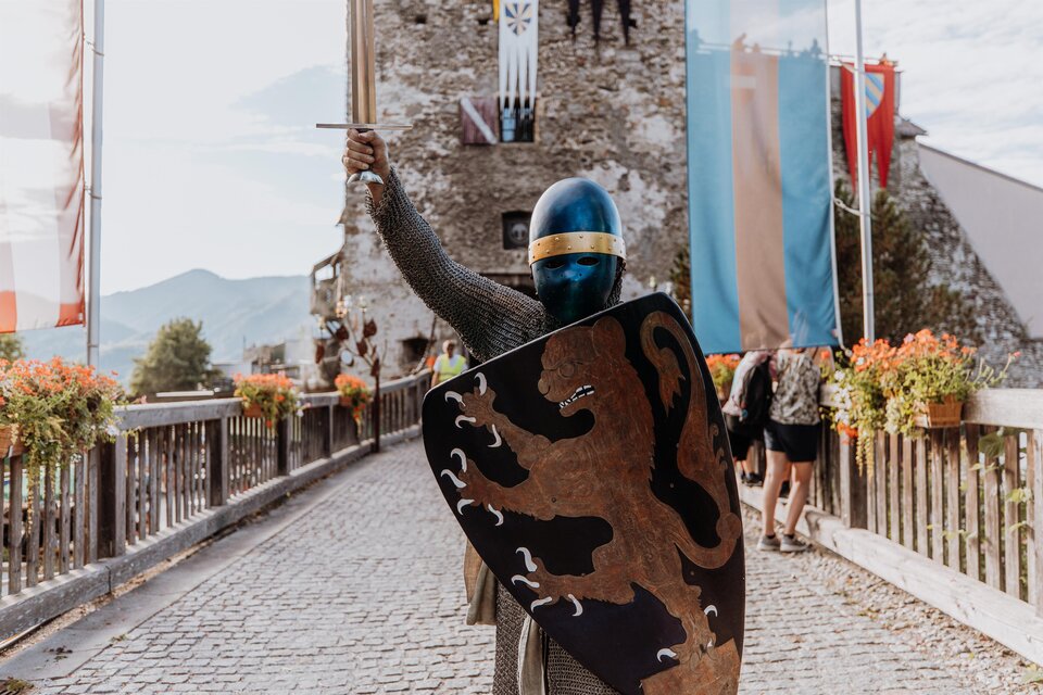 Ein Ritter in einer Rüstung steht auf einer Brücke vor einer Burg. Er hält ein Schwert in der Hand und zeigt ein Wappen auf seinem Schild. | © TV Hochsteiermark, nicoleseiser.at