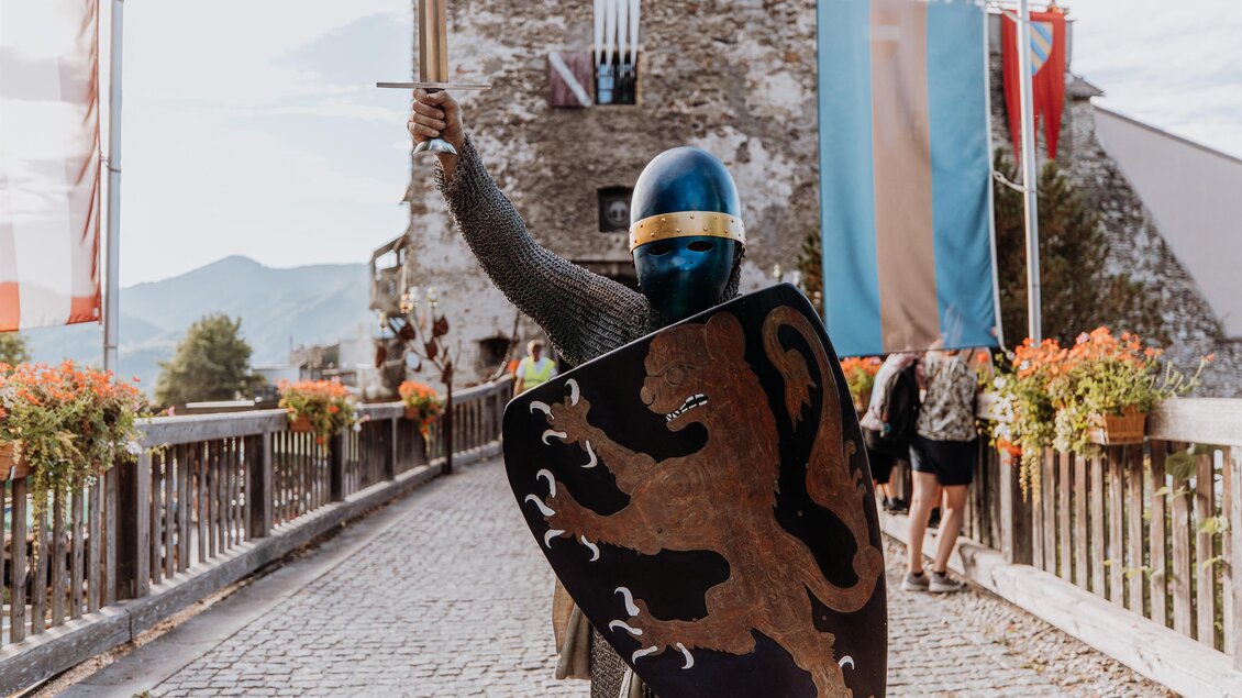 Ein Ritter in einer Rüstung steht auf einer Brücke vor einer Burg. Er hält ein Schwert in der Hand und zeigt ein Wappen auf seinem Schild. | © TV Hochsteiermark, nicoleseiser.at